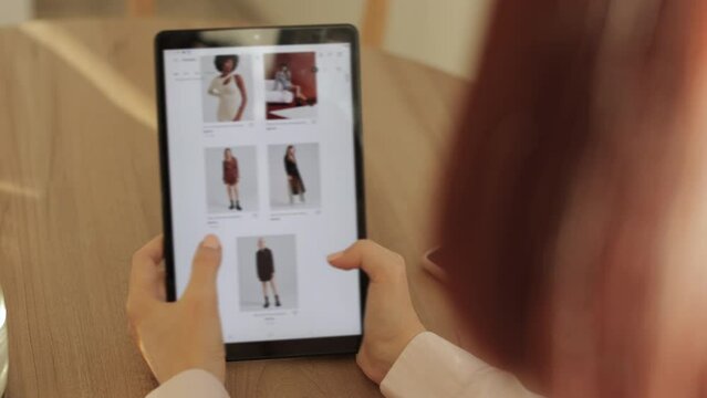 Rear view of female hand touching tablet screen while selecting dress on ecommerce portal. Lady use e-commerce webshop to buy dress. Modern devices, online, shopping concept