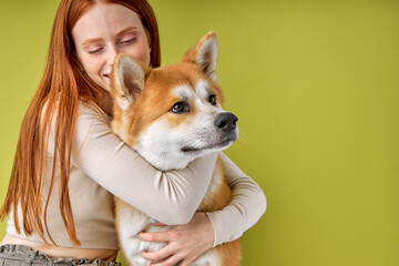 Red-haired lady in casual wear hugging purebred pet dog, akita inu dog with owner posing isolated over green studio background. Portrait copy space, for ad. animals, pets concept