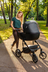 Young blonde woman walking with black stroller in summer park. Happy mother with baby in pram outdoors. 