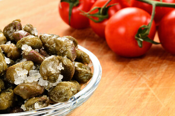 Salted capers in a bowl on a cutting board. Red tomatoes in the background.