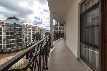 A wide curved balcony of a multi-storey building with metal black wrought iron railings with patterns. The balcony offers a view of the houses of the residential complex, mountains and clouds