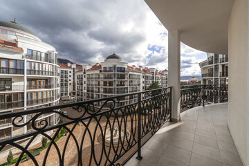 A wide curved balcony of a multi-storey building with metal black wrought iron railings with patterns. The balcony offers a view of the houses of the residential complex, mountains and clouds