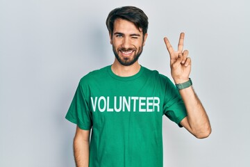 Young hispanic man wearing volunteer t shirt smiling with happy face winking at the camera doing...