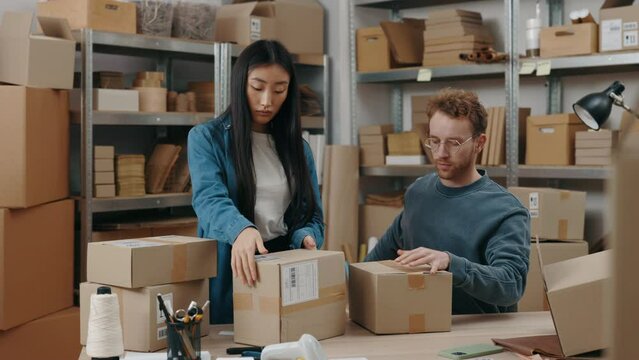 Established process. Two diverse male and female workers packing parcels while working together at the warehouse. Shelfs with cupboard boxes at the background. Small business concept.