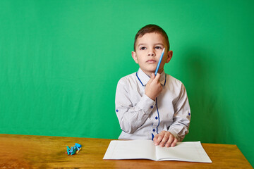 Cute pensive boy holding a pen in his hand sitting at a table with an open notebook on a green background.
