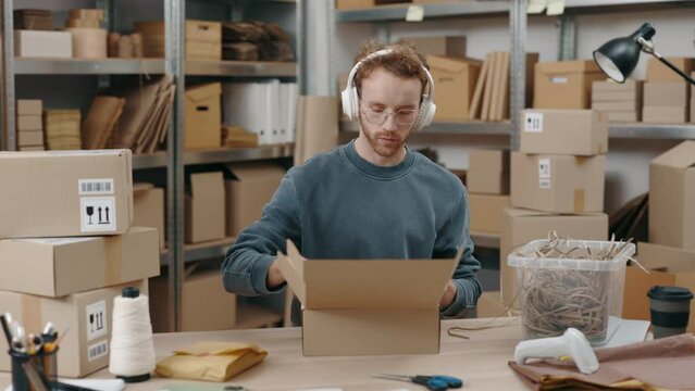 Waist up portrait view of the careful attentive man wearing headphones sitting at his workplace and putting filling at the box while packing parcels with light smile. Small business concept.