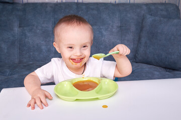 The child eats breakfast with a smile. The kid eats fruit puree. A little boy with down syndrome at...