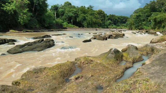 Muddy brown water of river flowing around big mossy rocks in tropical area.
