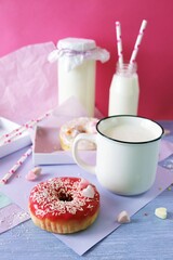 Freshly baked donuts glazed with various pink shades and sprinkled with caramel hearts, milk and yogurt in a cup and glass bottles, lilac-colored paper on the table