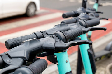 Simple electric scooters parked on the street, sidewalk, detail, handle bars closeup, nobody, zebra crossing, car in the background. Eco, green environmentally friendly urban city transport concept