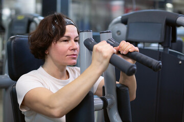 A middle-aged brunette woman is engaged in sports sitting on a simulator in the gym. A woman strengthens the muscles of the hands on the simulator.