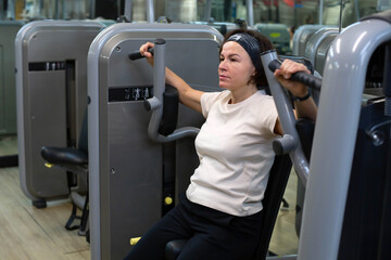 A middle-aged brunette woman is engaged in sports sitting on a simulator in the gym. A woman strengthens the muscles of the hands on the simulator.