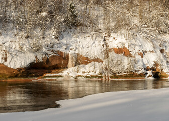 magical sunny winter day, landscape with red sandstone cliffs that are snowy with snow, frozen icicles on the cliff wall