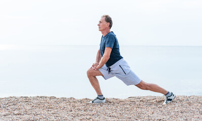 Active senior man is exercising outside. Elderly man is stretching legs early morning by sea beach. Healthy retirement lifestyle.