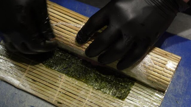 chef in kitchen in black sterile gloves spins long suchi roll in bamboo mat on blue board
