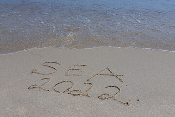 The inscription sea 2022 on the sand by the water and the rising wave, seashore beach vacation by the sea