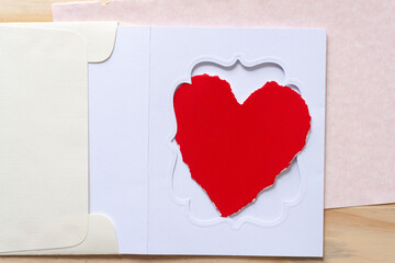 torn paper heart on a fancy windowed card inside and envelope on pink paper on a wooden surface
