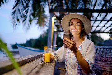 Vacation and technology. Young pretty woman in hat using smartphone sitting at beach cafe bar.