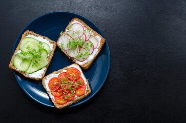appetizing delicious vegetarian sandwiches with cream cheese, fresh cucumber, radish, tomato and microgreens on a black slate background on a plate. Flat layout. healthy food diet weight loss fitness.