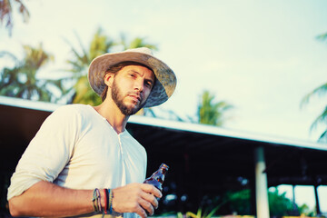 Vacation concept. Outdoor portrait of relaxed handsome young man with beard and hat holding bottle of beer at tropical resort.