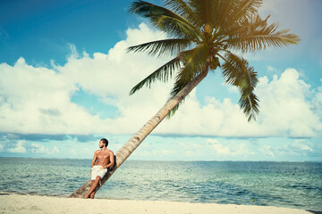 Enjoying suntan and vacation. Portrait of young bearded man leaning on coconut palm tree on the tropical  beach.