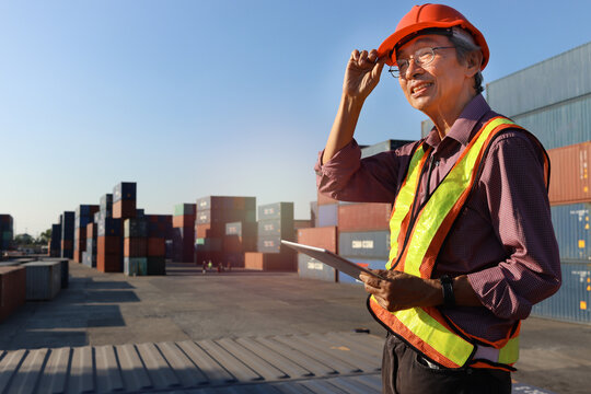 A senior elderly Asian worker engineer wearing safety vest and helmet standing and holding digital tablet at shipping cargo containers yard. elderly people at workplace concept