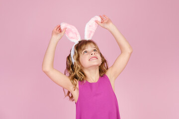 Easter concept - happy little caucasian girl raise head up touching bunny ears headband over pink background 
