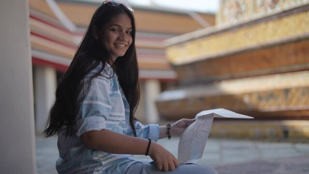 Asian woman use generic local map while sitting on pedestrian terrace. Young teen backpacker travel to sightseeing a landmark temple in Bangkok. Thailand.