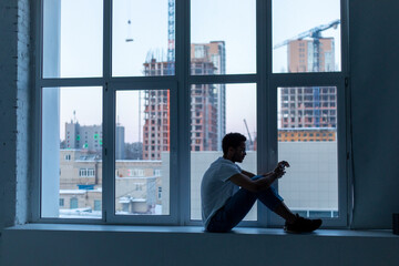 African American man in glasses and a white t-shirt uses a smartphone while sitting by the window.