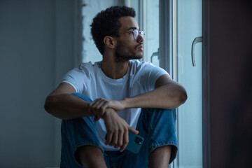 Attractive african american man in glasses and a white t-shirt sits on a windowsill.