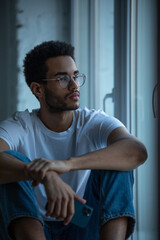 Fototapeta na wymiar Attractive african american man in glasses and a white t-shirt sits on a windowsill.