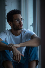 Fototapeta na wymiar Attractive african american man in glasses and a white t-shirt sits on a windowsill.