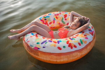 A smiling tween girl relaxing on a big donut shaped inflatable ring on a lake on a hot summer day,...