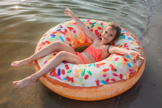 A smiling tween girl having fun on a big donut shaped inflatable ring on a lake on a hot summer day, happy summertime in countryside, local vacations, cottagecore