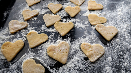 Raw dough in the form of hearts on a black glossy baking sheet sprinkled with flour