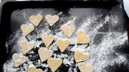 Raw dough in the form of hearts on a black glossy baking sheet sprinkled with flour