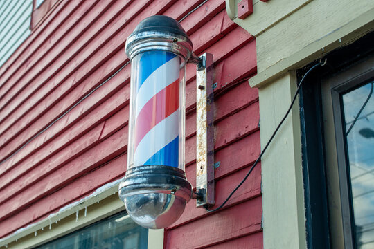 Barbershop pole with red, blue, and white stripes moving around on the outside of a barbershop. The vintage nostalgic pole is attached to a wall. The spiral pole is chrome with a glass cylinder case.