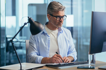 Successful senior boss with glasses, working on computer, businessman in shirt, in modern office at work