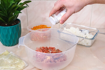 A man pours spices and salt into minced meat in a bowl for lazy cabbage rolls on the table.