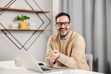 A successful entrepreneur sitting at his home office with a laptop on the desk and smiling at the...