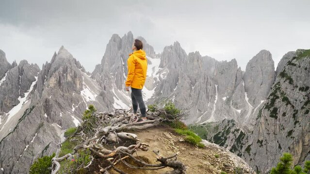 4K  Majestic rocky mountain peaks with woman in foreground wearing yellow jacket and enjoying landscape. Travelling, hiking, backpacking, exploring nature concept. Dolomite Alps, South Tyrol, Italy
