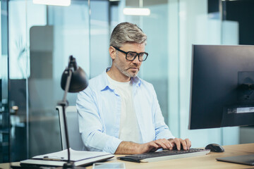 Successful senior boss with glasses, working on computer, businessman in shirt, in modern office at...