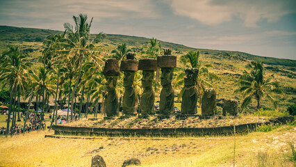 7 Moais mit roten Hüten, den Pukaus, stehen vor Palmen am paradiesischen Strand von Playa Anakena auf Rapa Nui der Osterinsel.
