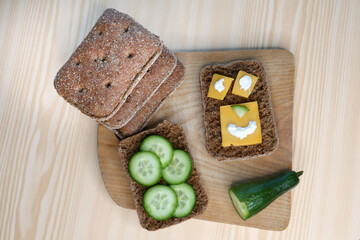 Happy snacks. Breakfast nutrition, food on a wooden cutting board placed on the table. Healthy diet making with rye cereal bread, cheese and cucumbers combination.