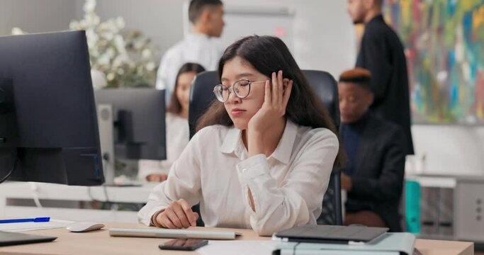Bored pretty office secretary employee in a white shirt sits in front of the computer monitor, she is tired, wants to sleep, supports her face with hand, waiting for the end of work