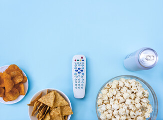 TV remote control, popcorn, can of beer, nachos and chips on light blue background. Сoncept of family watching movies and television. Selective focus, copy space