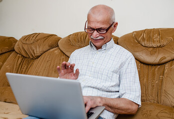 cheerful old man 50s rests on a comfortable sofa and communicates with relatives on a laptop. An elderly man remotely talks and waves to his grandchildren who called online on a computer gadget.