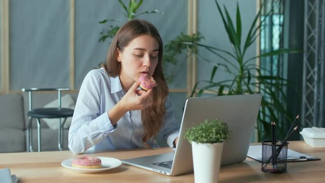 Young Businesswoman Eating a Glazed Donut While Working on a Laptop at the Office Table. A Quick Snack, High-calorie Food, Weight Problems, Diabetes Mellitus. Eating Disorder and Business Concept.