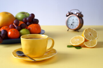 Healthy breakfast of fruits and tea with lemon. There is a yellow tablecloth on the table, a cup of tea is yellow. There is a blurry alarm clock in the photo that shows 7 o'clock.