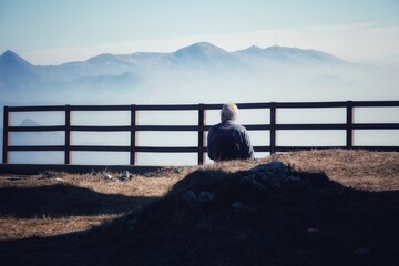Persona anziana di spalle con camicia a quadri che osserva il paesaggio seduto in montagna - obrazy, fototapety, plakaty